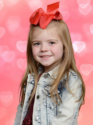 Young girl smiling, wearing a jean jacket and big red hairbow, in front of a pink heart backdrop