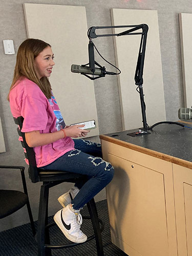 Teenage girl sitting on a stool next to a professional recording microphone in a soundbooth
