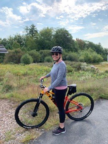 woman wearing a helmet and sunglasses, standing next to a bicycle on a rural trail