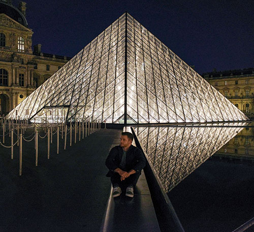 man sitting on stone path in front of illuminated Louvre Museum pyramid at night