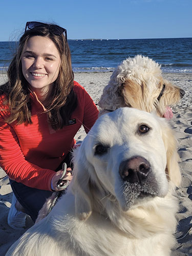 smiling young woman, crouched next to two light-colored dogs at the beach
