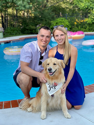 smiling young man and woman crouched next to light-colored dog in front of a blue pool