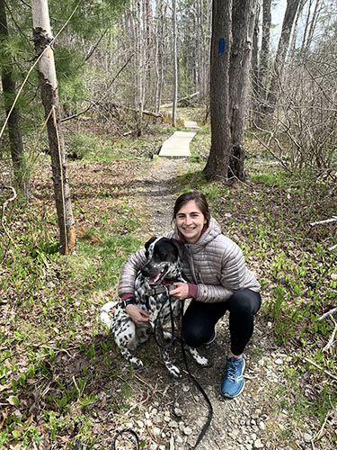Young woman crouched next to a black and white dog, together on a wooded walking trail
