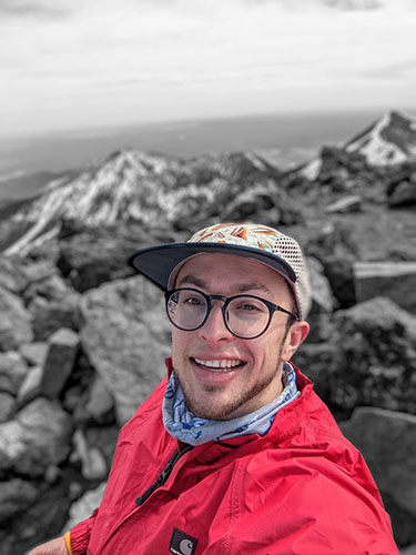 Young man in a white hat, glasses, and red jacket in front of a rocky coastline