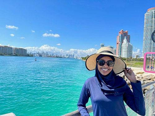 woman in a sunhat and glasses in front of green, warm sea water. Large buildings are in the backdrop.