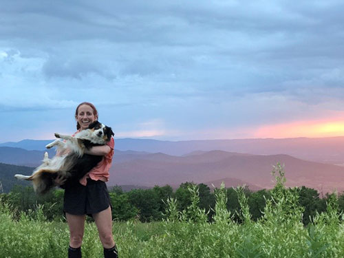 Woman holding a dog with a mountain range and sunset behind her