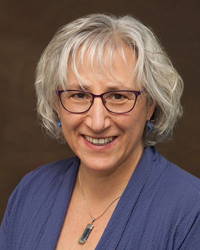 Susan Gatti, smiling woman with white/silver hair and glasses wearing a blue shirt ad silver necklace