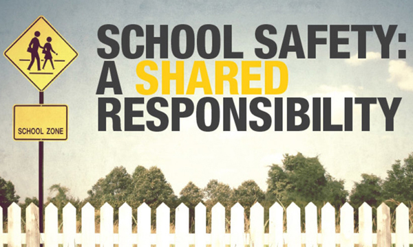 Crosswalk sign in front of white-picket fence with text "School Safety: A Shared Responsibility"