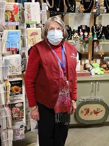 older woman wearing a red vest and surgical mask, standing in a gift shop