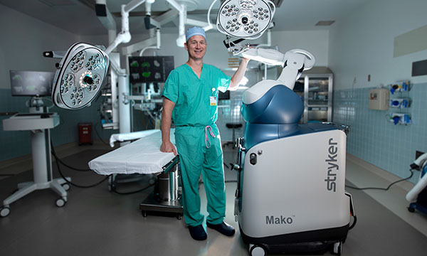 Dr. Jeff Bush wearing scrubs and standing next to Mako robot in a surgical suite