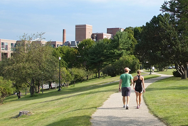 Two people walk on a path surrounded by green grass and trees. Maine Medical Center is seen in the distance.
